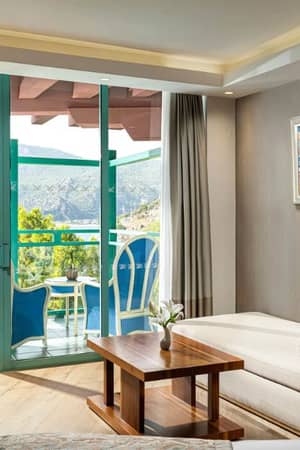 Hotel room at Liberty Lykia Resort with a comfortable lounge area, balcony with blue chairs, and a view of greenery and mountains.