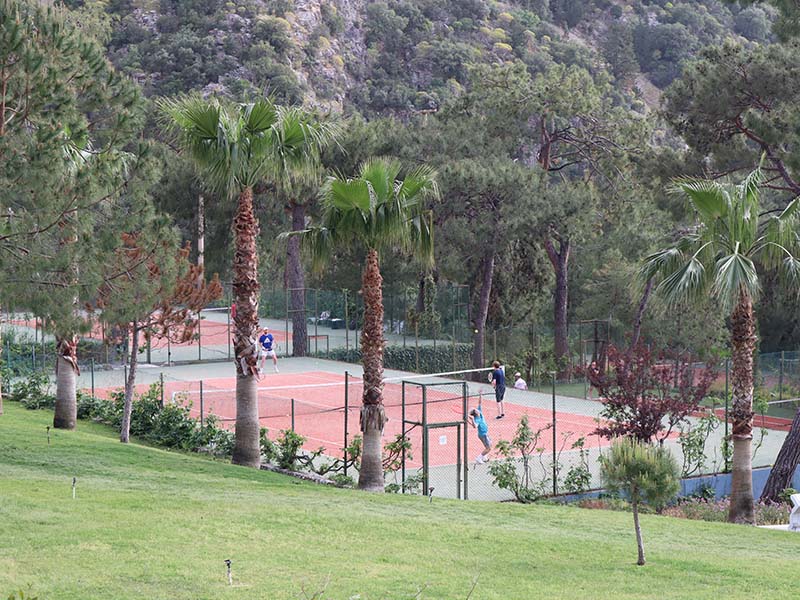 Tennis players on clay courts at Liberty Lykia Tennis Academy, surrounded by lush palm trees and hills.