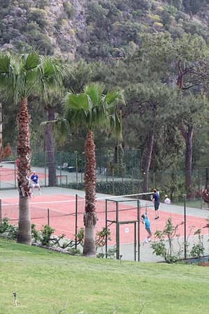 Tennis players on clay courts at Liberty Lykia Tennis Academy, surrounded by lush palm trees and hills.