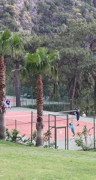 Tennis players on clay courts at Liberty Lykia Tennis Academy, surrounded by lush palm trees and hills.