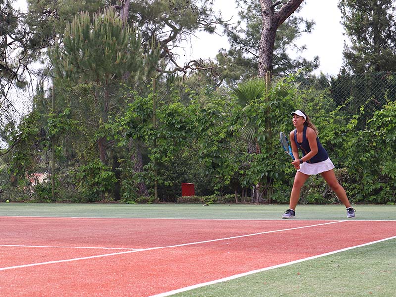Woman standing ready on a tennis court at Liberty Lykia Tennis Academy, surrounded by trees and greenery.