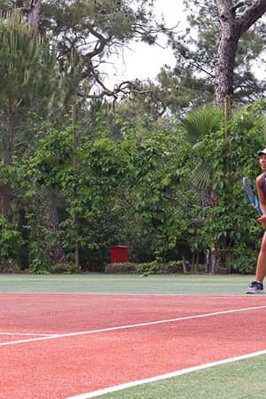 Woman standing ready on a tennis court at Liberty Lykia Tennis Academy, surrounded by trees and greenery.