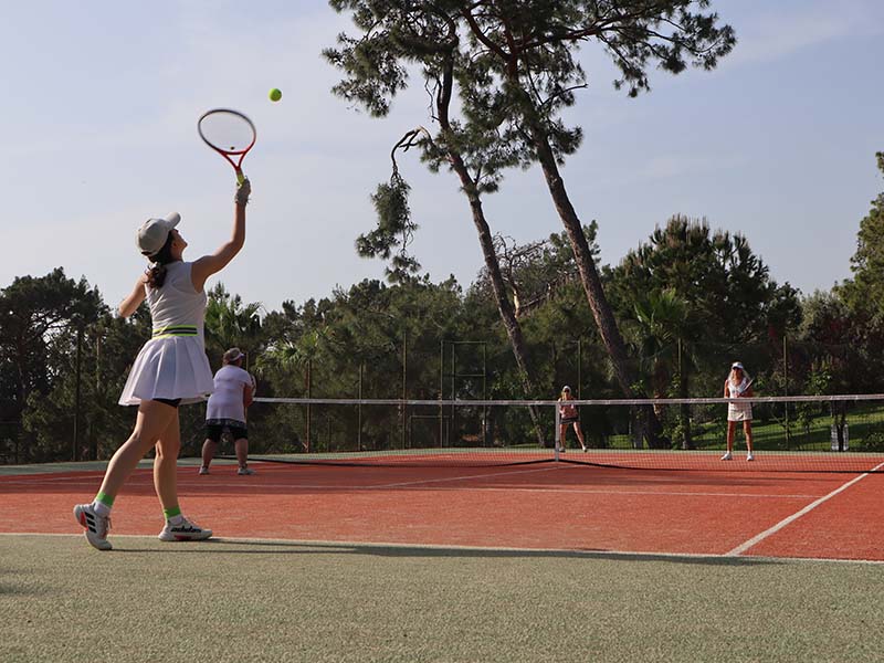 Outdoor tennis match at Liberty Lykia Tennis Academy with a woman serving on a court surrounded by trees.
