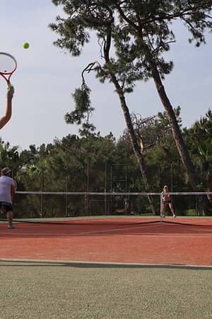 Outdoor tennis match at Liberty Lykia Tennis Academy with a woman serving on a court surrounded by trees.