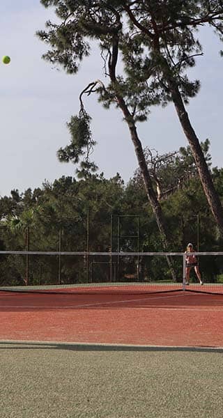 Outdoor tennis match at Liberty Lykia Tennis Academy with a woman serving on a court surrounded by trees.