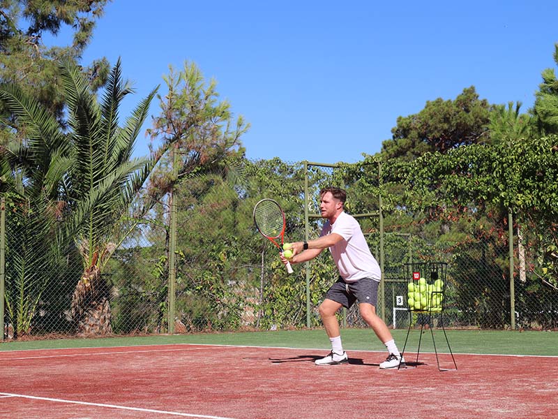 Tennis player preparing to hit a ball on a clay court at Liberty Lykia Tennis Academy, with palm trees and a tennis ball basket visible.