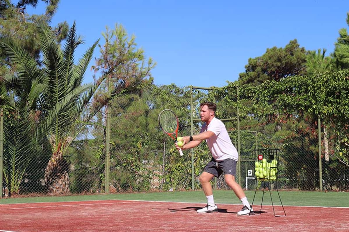 Tennis player preparing to hit a ball on a clay court at Liberty Lykia Tennis Academy, with palm trees and a tennis ball basket visible.