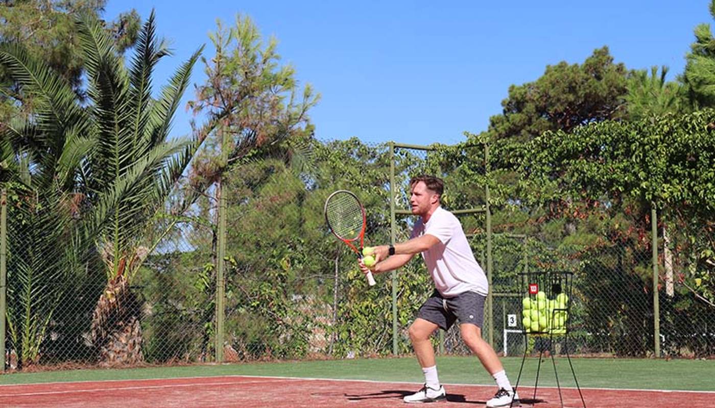Tennis player preparing to hit a ball on a clay court at Liberty Lykia Tennis Academy, with palm trees and a tennis ball basket visible.