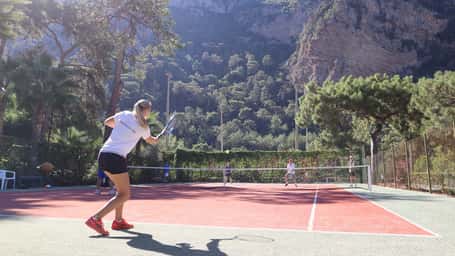 Outdoor tennis match in progress at Liberty Lykia Tennis Academy with cliffs and greenery in the background.