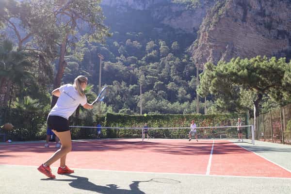 Outdoor tennis match in progress at Liberty Lykia Tennis Academy with cliffs and greenery in the background.