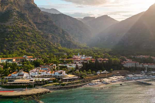 Aerial view of Liberty Lykia Village resort on the Mediterranean coast, showing buildings, green mountains, and a sandy beach.