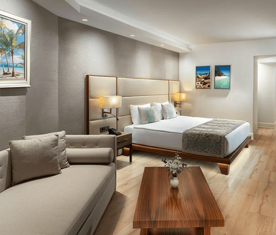 Luxurious hotel room with king-sized bed, beige sofa, wooden table, and seascape artwork on the walls, under warm ambient lighting.