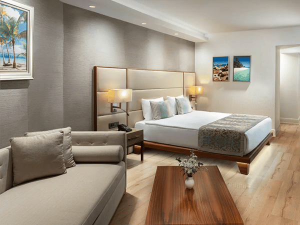 Luxurious hotel room with king-sized bed, beige sofa, wooden table, and seascape artwork on the walls, under warm ambient lighting.