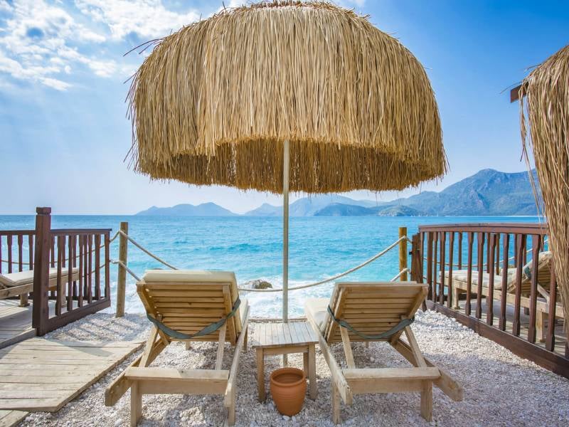 Two loungers on a pebble beach under a straw parasol, facing turquoise sea and mountains at Liberty Sentido Gallery.