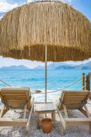 Two loungers on a pebble beach under a straw parasol, facing turquoise sea and mountains at Liberty Sentido Gallery.