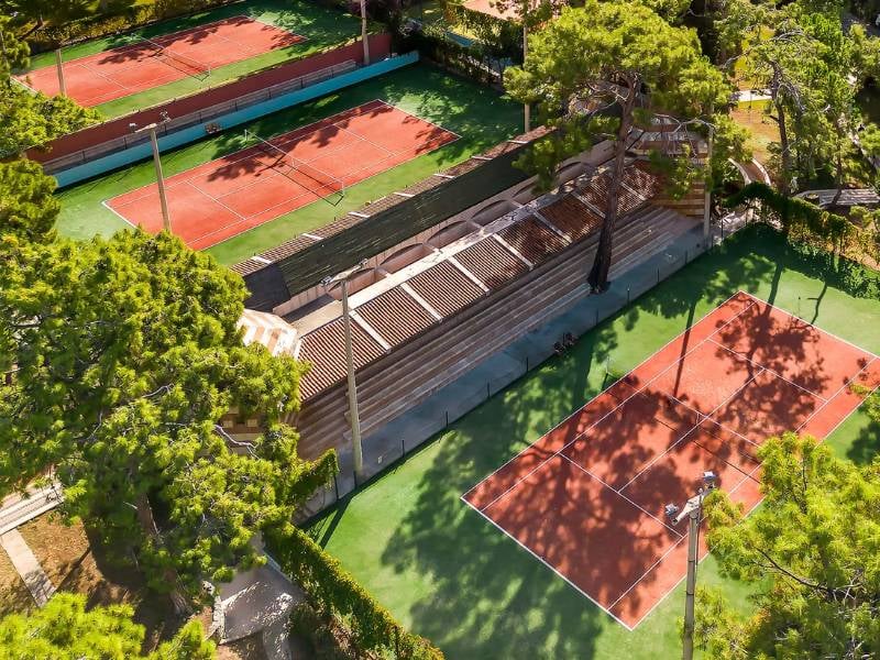 Aerial view of tennis courts among lush trees with players on clay surfaces.