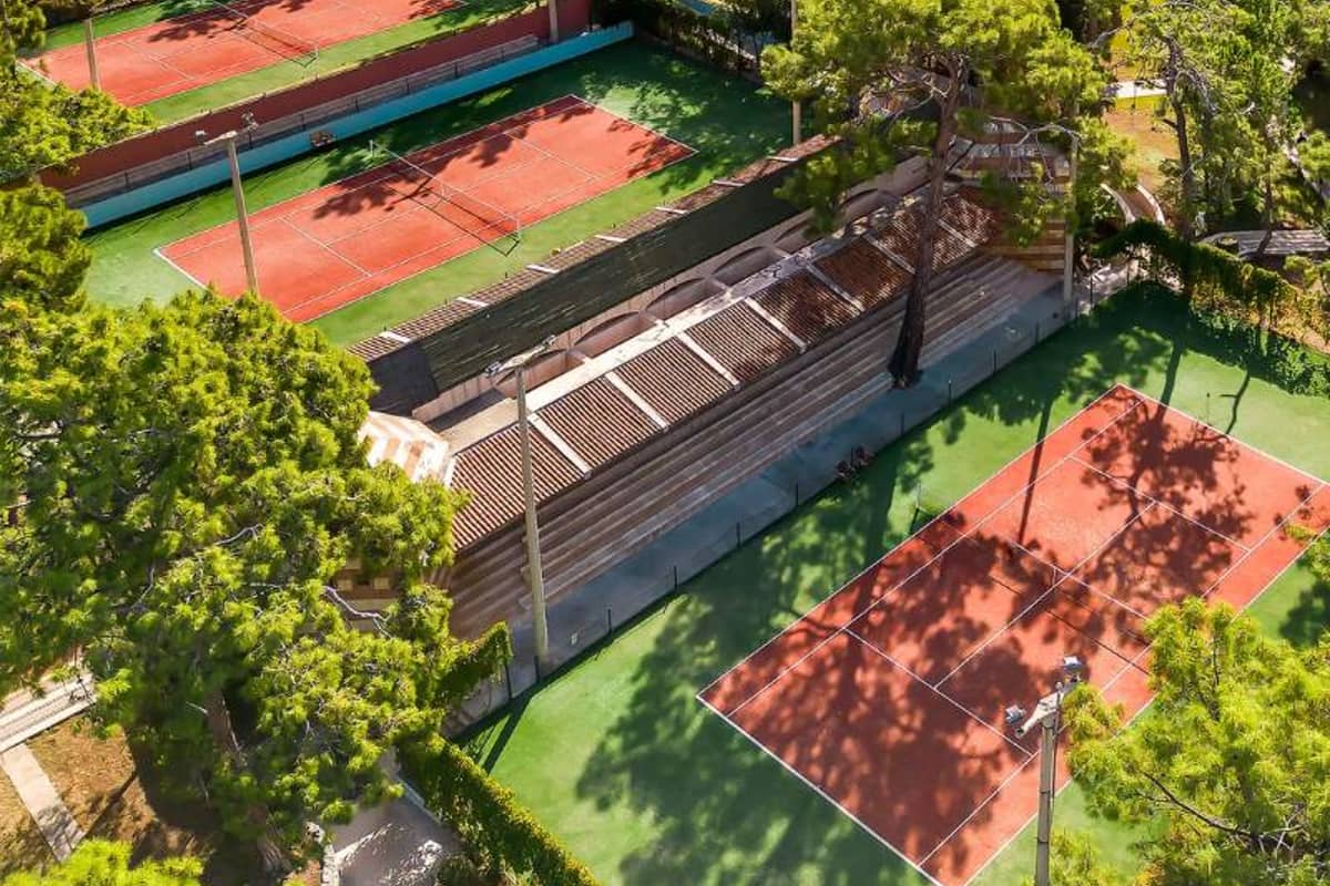 Aerial view of tennis courts among lush trees with players on clay surfaces.