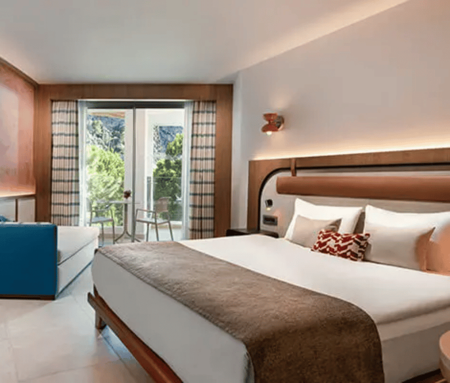 Liberty Village deluxe room with a side sea view, featuring a double bed, blue sofa, and balcony overlooking greenery and sea.