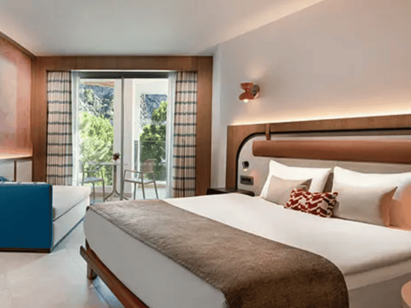 Liberty Village deluxe room with a side sea view, featuring a double bed, blue sofa, and balcony overlooking greenery and sea.