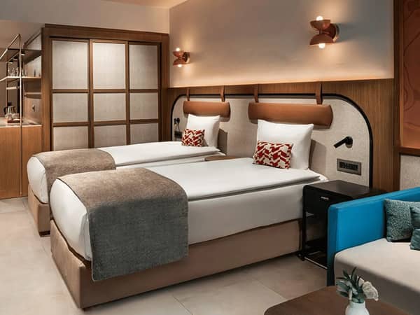 Liberty Village FTH Deluxe Family Room showcasing two single beds, decor featuring wood panels, a blue sofa, and a view of the sea from the side.