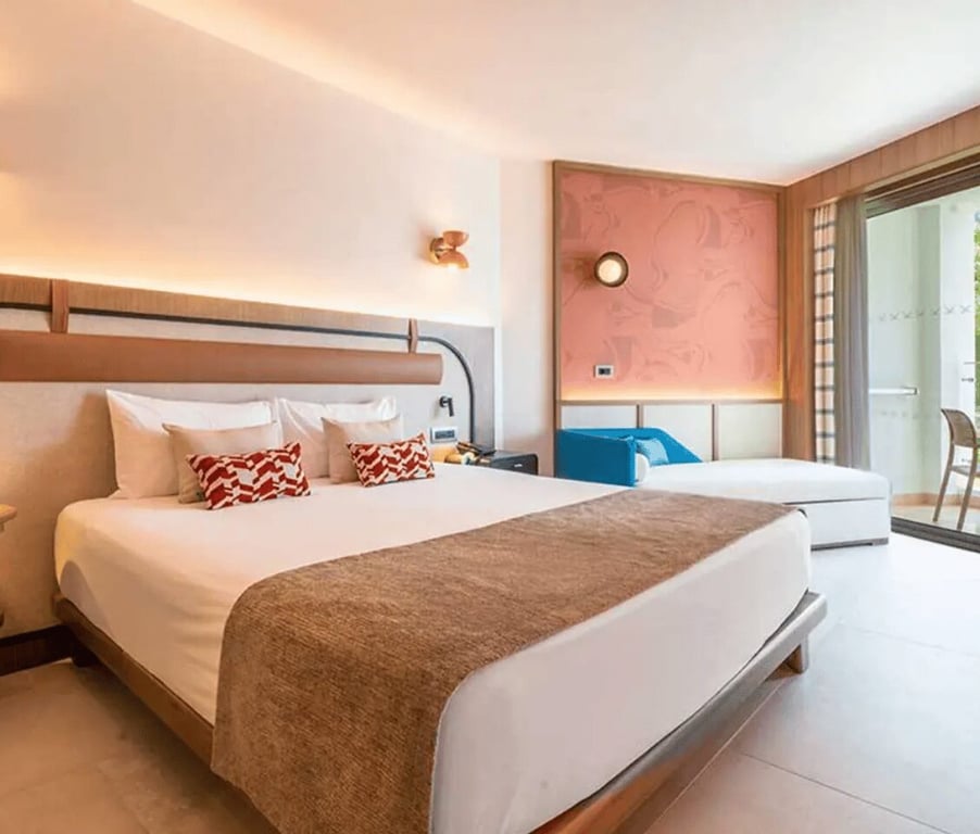 Interior of a Deluxe Standard Land Side hotel room at Liberty Village, with a king-sized bed, decorative pillows, and a balcony with outdoor seating.