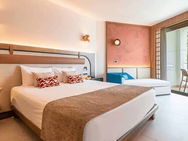 Interior of a Deluxe Standard Land Side hotel room at Liberty Village, with a king-sized bed, decorative pillows, and a balcony with outdoor seating.