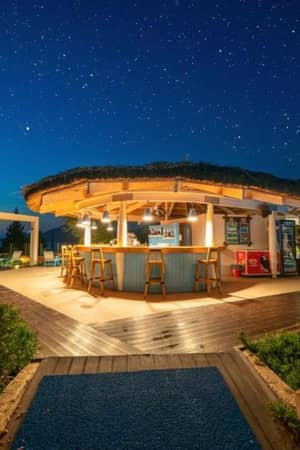 Night bar at Liberty Village Gallery with a round thatched roof, wooden stools, surrounded by greenery, and a star-filled sky.