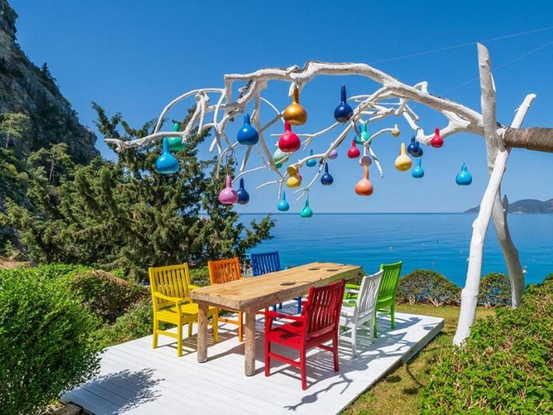 Outdoor seating area at Liberty Village Gallery with a wooden table, colourful chairs, and a tree sculpture decorated with multi-coloured lights, overlooking the sea.