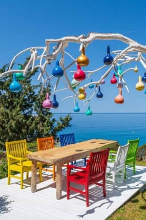Outdoor seating area at Liberty Village Gallery with a wooden table, colourful chairs, and a tree sculpture decorated with multi-coloured lights, overlooking the sea.