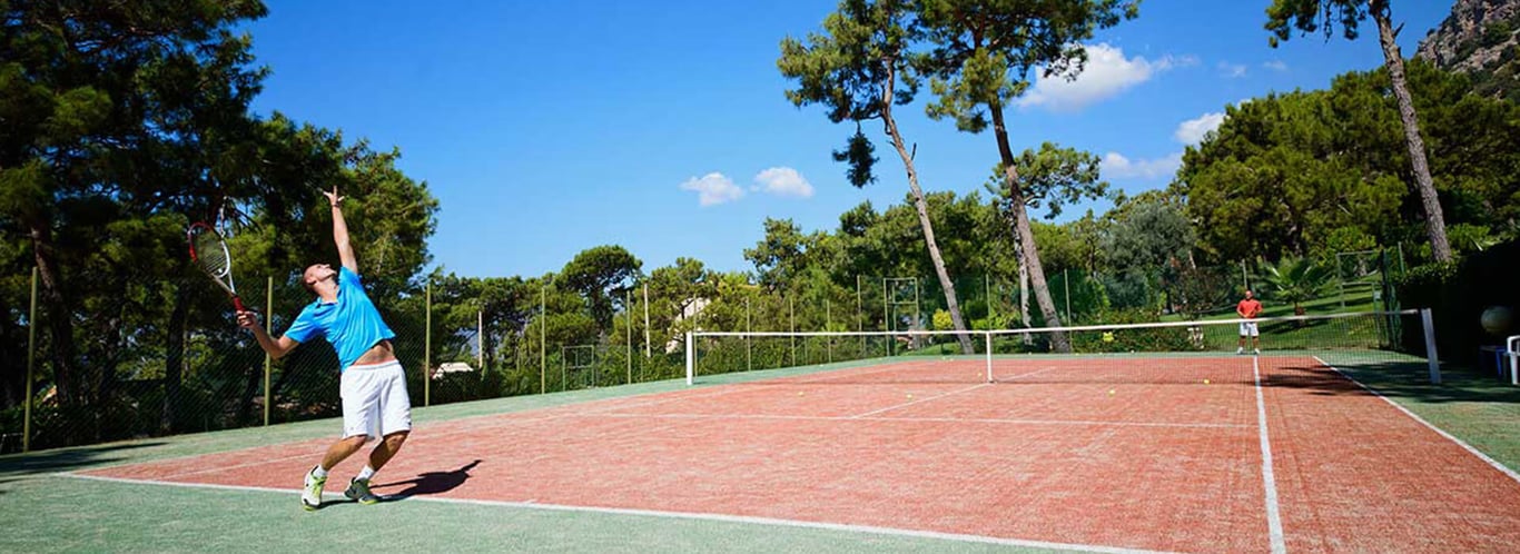 Outdoor tennis court at Liberty Village with a male player in blue shirt serving and another player waiting, surrounded by greenery and tall trees.