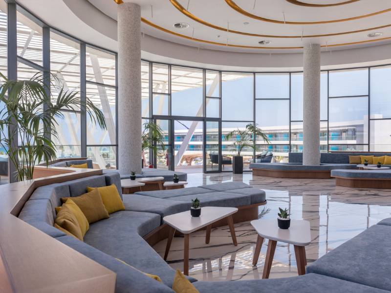 Modern lounge area at Lyttos Gallery with grey sofas, yellow cushions, white tables, and floor-to-ceiling windows with an outdoor view.