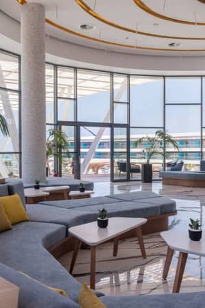 Modern lounge area at Lyttos Gallery with grey sofas, yellow cushions, white tables, and floor-to-ceiling windows with an outdoor view.