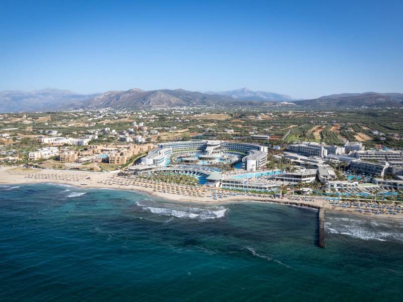 Aerial view of Lyttos Beach Resort in Crete, Greece, showcasing pools, beach, and mountainous backdrop.