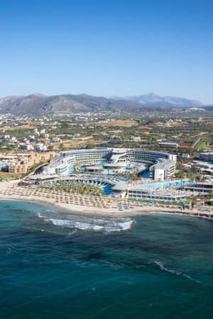 Aerial view of Lyttos Beach Resort in Crete, Greece, showcasing pools, beach, and mountainous backdrop.