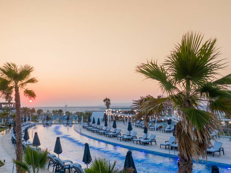 Aerial view of Lyttos Beach hotel pool area at sunset, with an infinity pool, sun loungers, and palm trees.