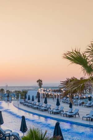 Aerial view of Lyttos Beach hotel pool area at sunset, with an infinity pool, sun loungers, and palm trees.