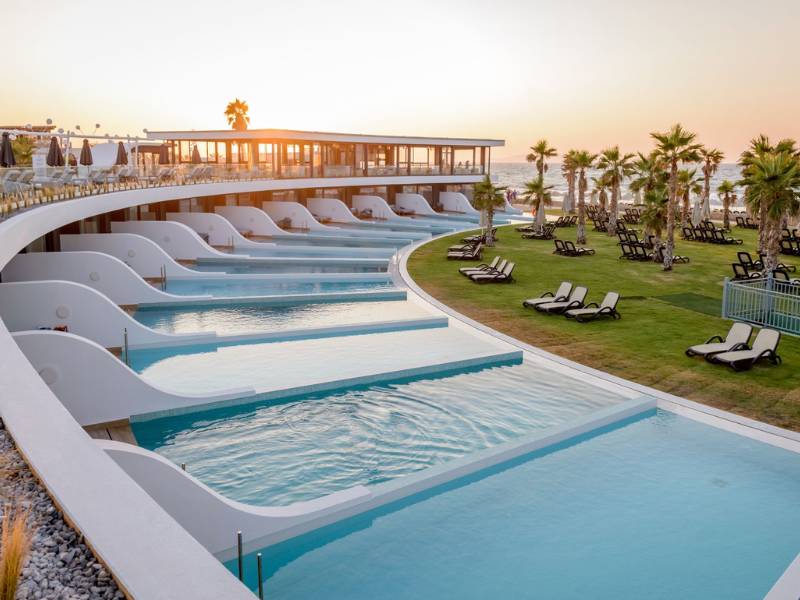 Lyttos Gallery resort pool area at sunset with infinity pools, sun loungers, palm trees, and ocean view.