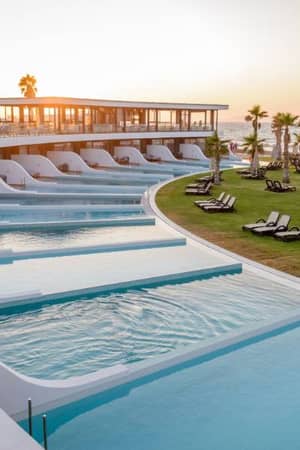 Lyttos Gallery resort pool area at sunset with infinity pools, sun loungers, palm trees, and ocean view.