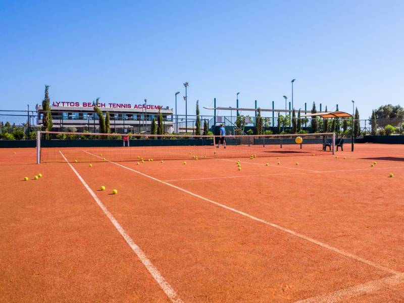 Clay tennis court at Lyttos Beach Tennis Academy with tennis balls scattered on the ground and a net in the centre.