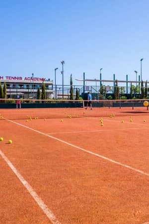 Clay tennis court at Lyttos Beach Tennis Academy with tennis balls scattered on the ground and a net in the centre.