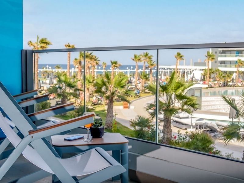 Balcony view from Lyttos Mare Resort with chairs, table, palm trees, pool, and sea.