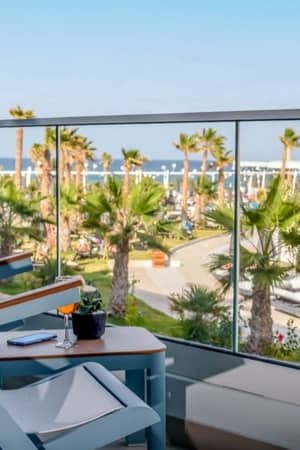 Balcony view from Lyttos Mare Resort with chairs, table, palm trees, pool, and sea.