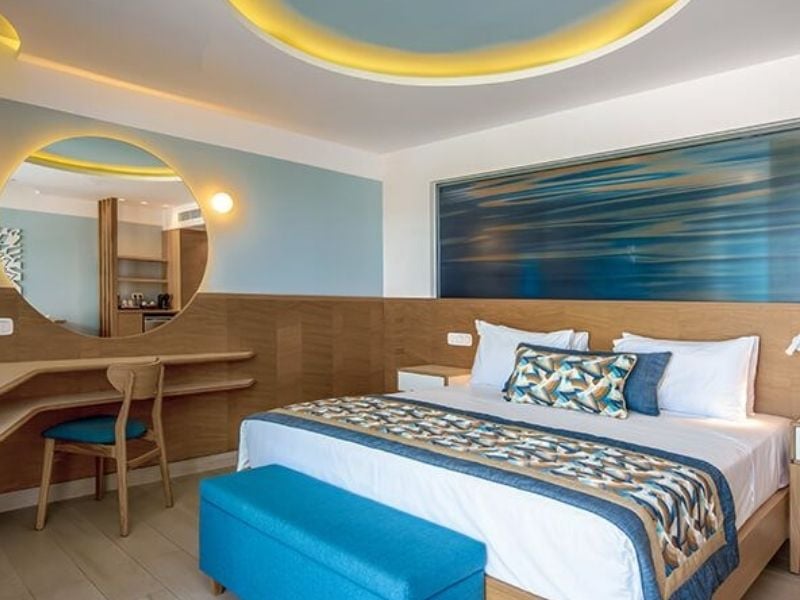 Hotel room at Lyttos Beach with a large bed, blue and beige linens, a wood desk with a round mirror, and ambient lighting.