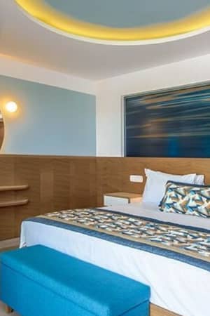 Hotel room at Lyttos Beach with a large bed, blue and beige linens, a wood desk with a round mirror, and ambient lighting.
