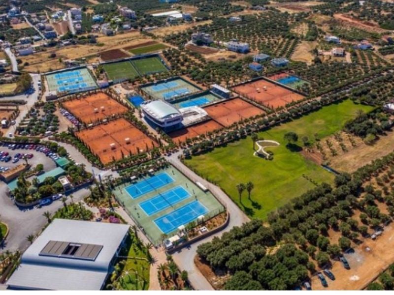Aerial view of the Lyttos Mare tennis complex with multiple clay and hard tennis courts, olive groves, and a central building.