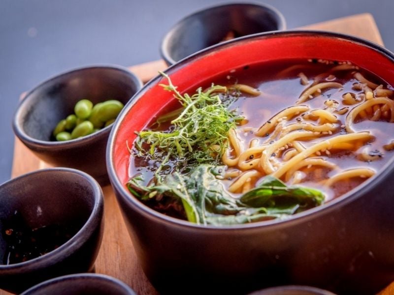 Asian noodle soup in a black and red bowl with herbs, surrounded by bowls of edamame and condiments on a wooden tray.