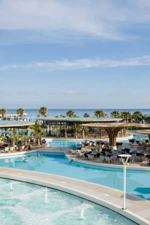 Lyttos Mare resort with luxurious turquoise pools, sun loungers, palm trees, and a sea view in the background.