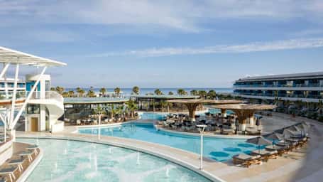 Lyttos Mare resort with luxurious turquoise pools, sun loungers, palm trees, and a sea view in the background.