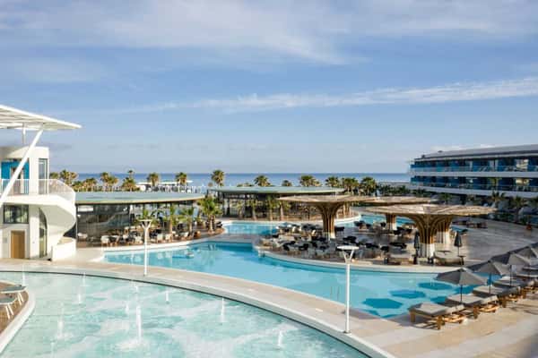 Lyttos Mare resort with luxurious turquoise pools, sun loungers, palm trees, and a sea view in the background.