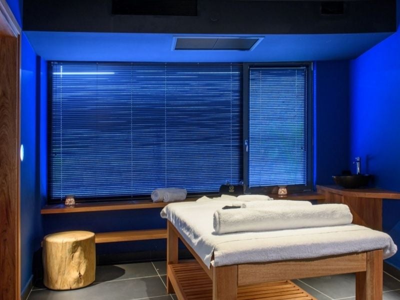 Lyttos Mare spa room with massage table, wooden bench, candles, and deep blue walls for a calming atmosphere.